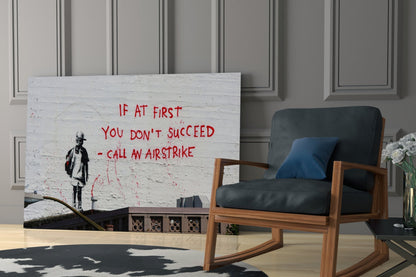 Glass art print displaying red text reading "IF AT FIRST YOU DON'T SUCCEED – CALL AN AIRSTRIKE" with a stenciled figure, displayed in a modern living room next to a wooden chair.
