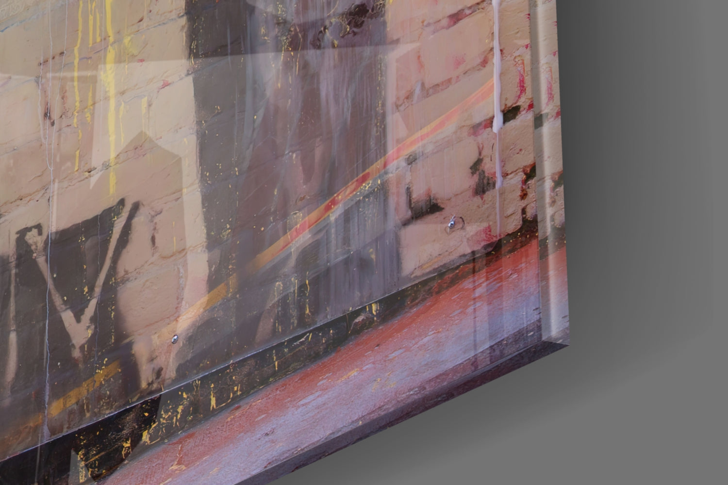 Angled close-up of transparent glass art panel showing layered urban imagery with red, yellow, and brown tones against gray wall.