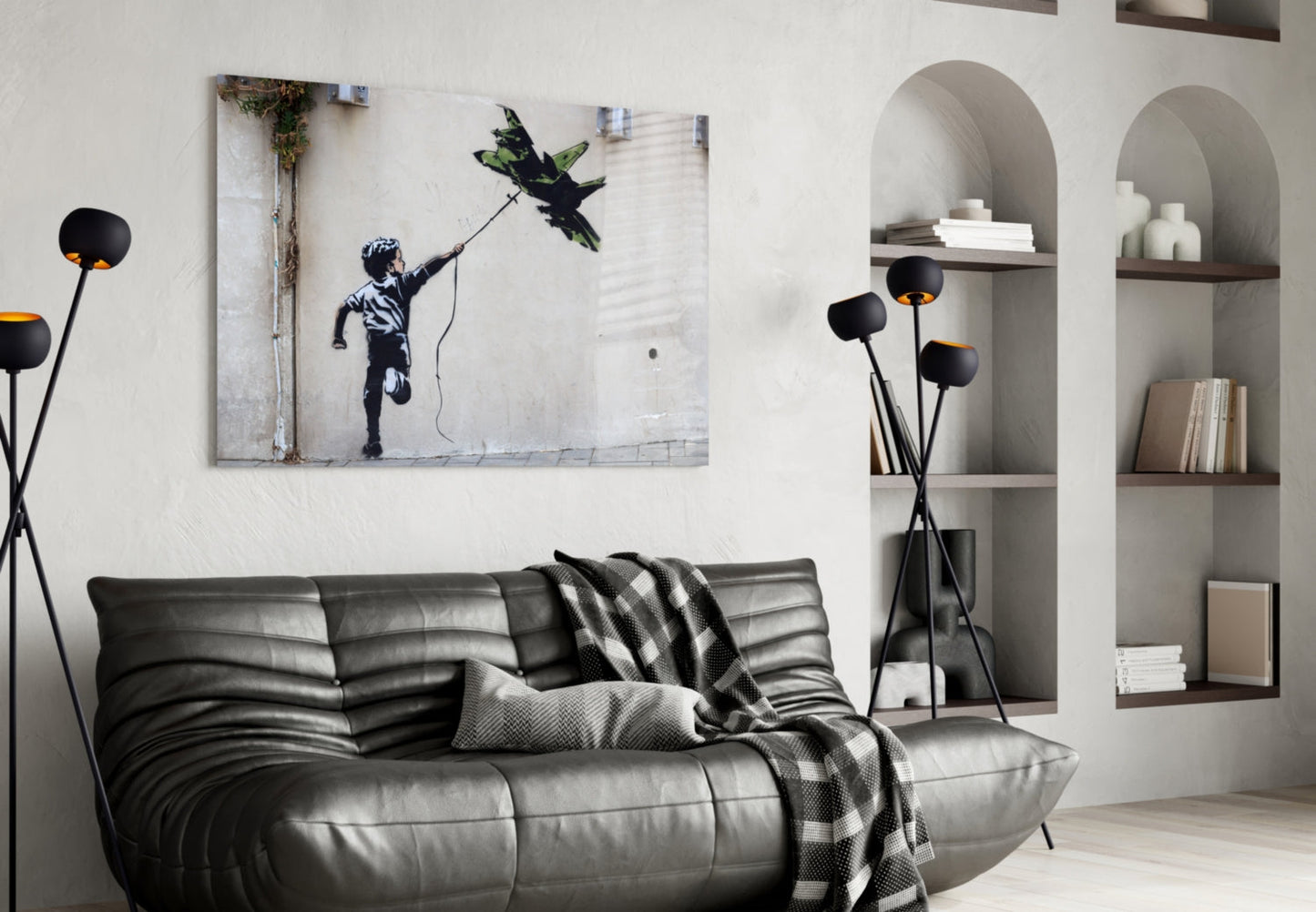 Black leather sofa with patterned throw blanket beneath framed Banksy-style street art print of child reaching for kite, flanked by arched wall shelving.