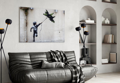 Black leather sofa with patterned throw blanket beneath framed Banksy-style street art print of child reaching for kite, flanked by arched wall shelving.