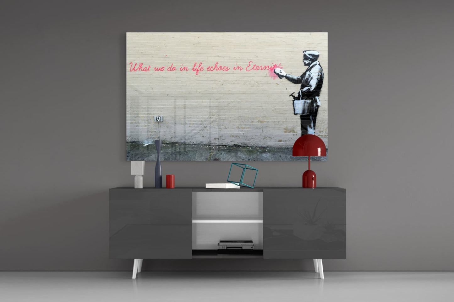Banksy-style street art glass print mounted above gray media console displaying stenciled figure with red text quote and red decorative accessories.