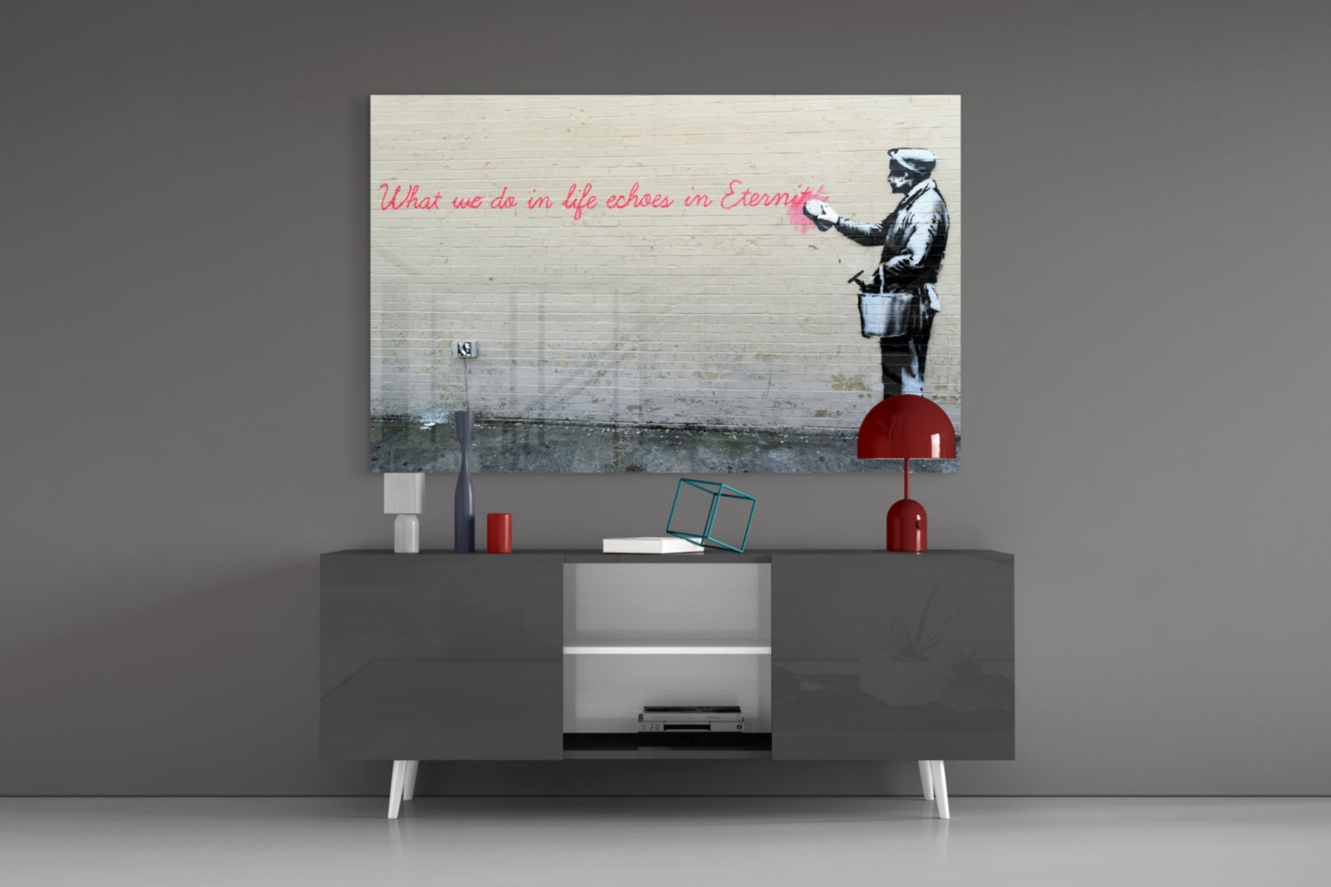 Banksy-style street art glass print mounted above gray media console displaying stenciled figure with red text quote and red decorative accessories.