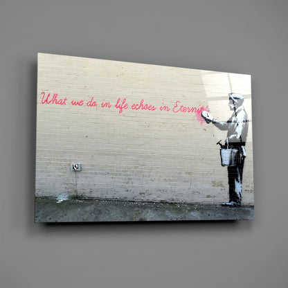 Glass art print displayed on gray wall showing stenciled figure in white with pink handwritten text on white brick background.