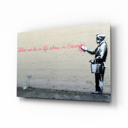 Glass art print displayed at an angle showing stenciled figure with bucket and pink handwritten text on wooden wall background.
