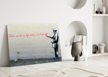 Glass art print leaning against white wall displaying Banksy-style stenciled figure with red handwritten text in minimalist room setting.