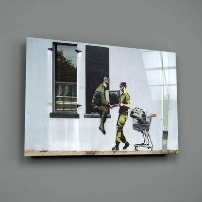 Glass art print depicting two olive-uniformed soldiers exchanging a television and shopping cart against white brick wall with windows.