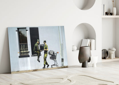 Glass art print leaning against white wall showing two figures in yellow-green carrying television and shopping cart.