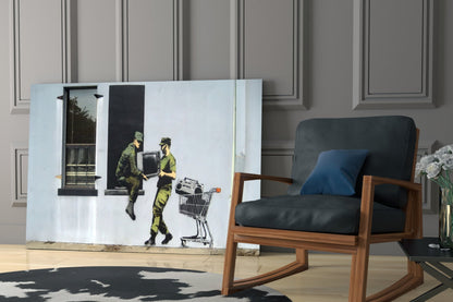 Soldier Glass Art depicting two figures in military camouflage with shopping cart displayed against paneled wall beside wooden chair.