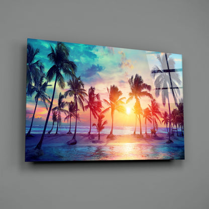 Glass art panel mounted on gray wall displaying tropical beach sunset with palm trees, golden sun, and colorful sky reflections.