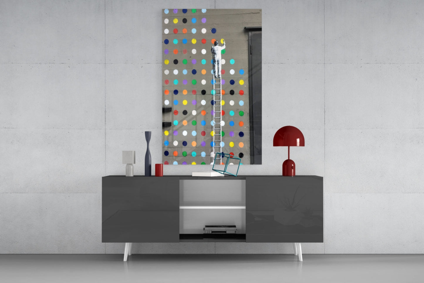 Artist on ladder installing colorful glass dots panel above gray media console in modern interior setting.