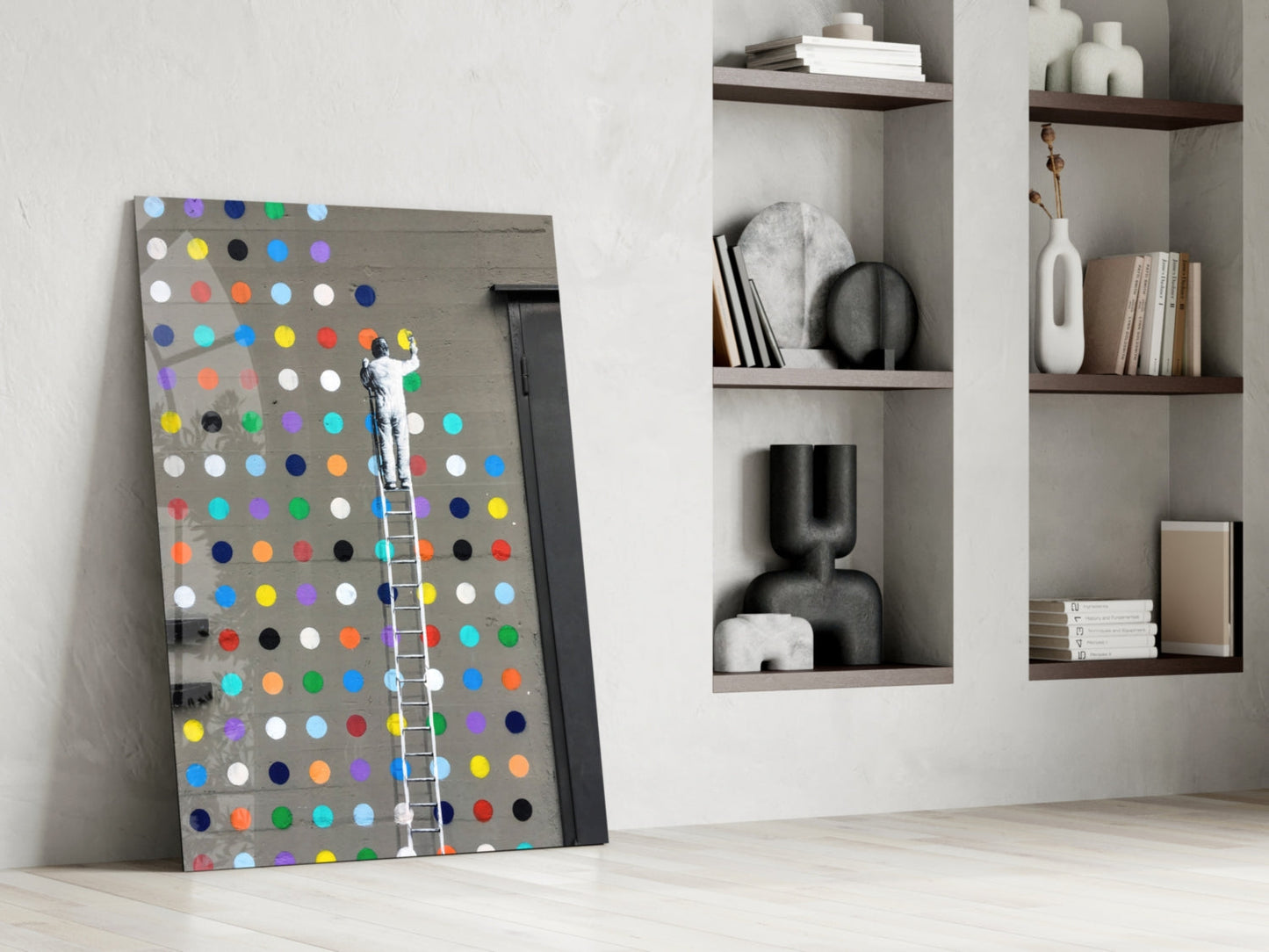 Colorful dots glass art leaning against white wall with figure on ladder, beside styled white shelving with books and decorative objects.
