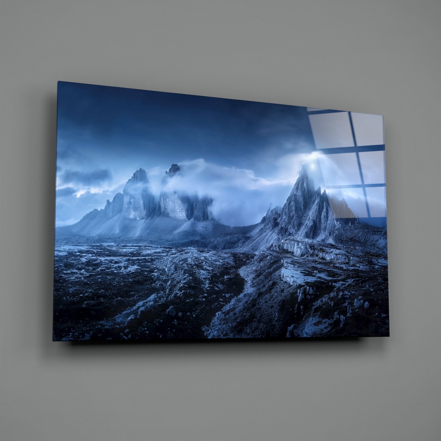 Mounted glass art panel displaying dramatic blue-toned mountain peaks with snow and cloudy sky, mounted on gray wall.