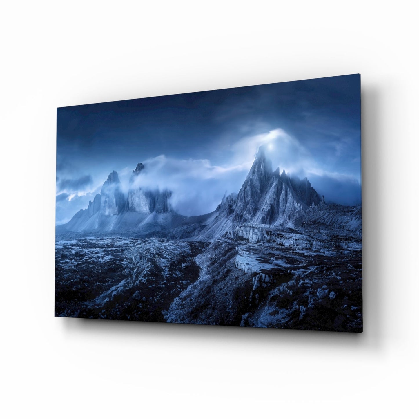 Angled perspective view of glass art panel displaying dramatic blue-toned mountain landscape with jagged peaks and cloudy sky, mounted on white background.