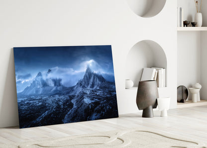 Glass art panel leaning against white wall displays snow-covered jagged mountains under dramatic blue sky with light rays.
