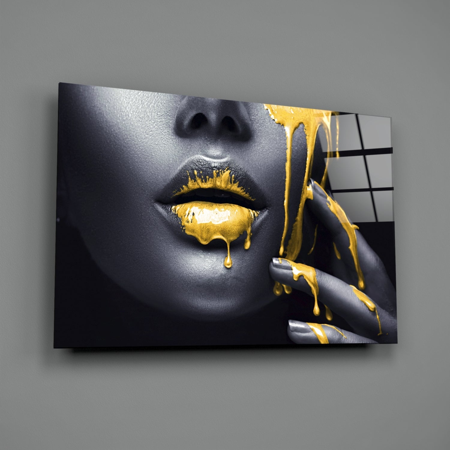 Glass wall art displaying a woman's face with gray skin tone and gold dripping paint effects on lips and fingers, mounted on gray wall.