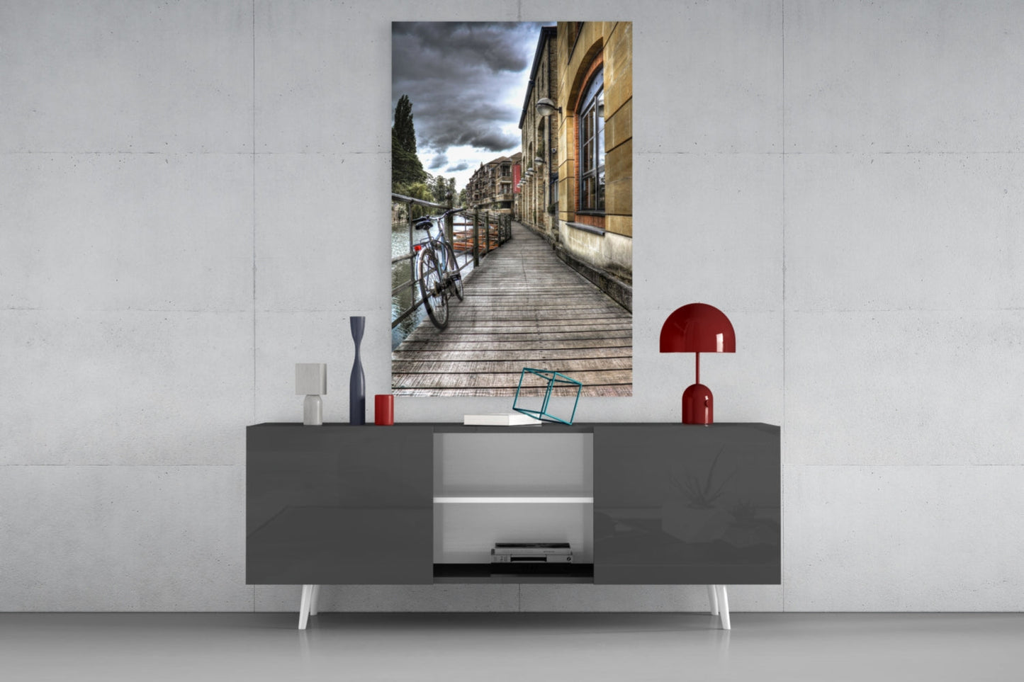 Vertical glass wall art depicting a wooden bridge alongside historic buildings with a bicycle, displayed above a gray media console with red lamp and decorative objects.
