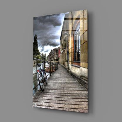 Wooden boardwalk alongside historic stone buildings with bicycle parked at railing under dramatic cloudy sky, displayed as angled glass wall art.