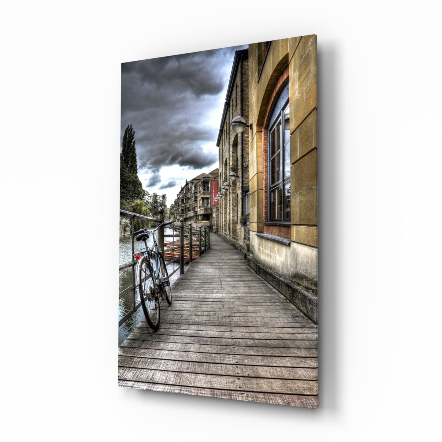 Bicycle parked on wooden boardwalk beside stone building with dramatic cloudy sky and tree-lined canal in background.