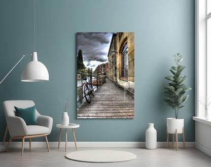 Wooden waterside boardwalk with red bicycle, stone building, and dramatic cloudy sky displayed as framed glass wall art in modern room setting.