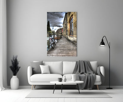 Wooden boardwalk along stone building with parked bicycle and cloudy sky, displayed as glass wall art above living room sofa.