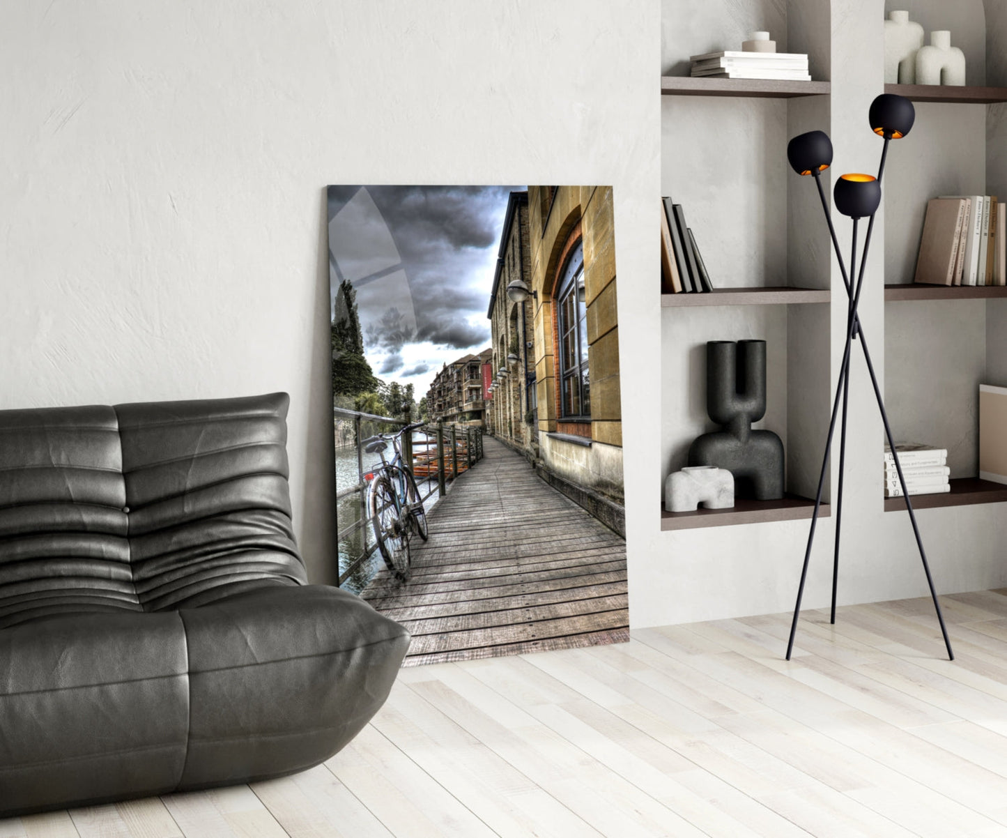 Modern living room with glass wall art depicting a waterside street scene with bicycle, displayed between gray leather furniture and white shelving with black decorative pieces.