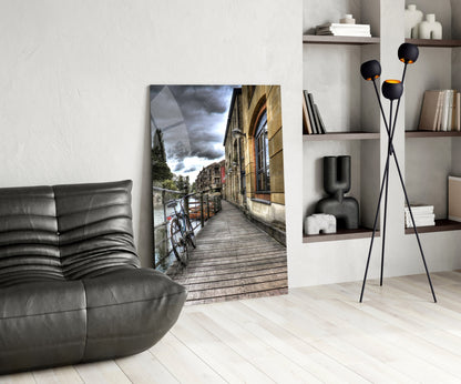 Modern living room with glass wall art depicting a waterside street scene with bicycle, displayed between gray leather furniture and white shelving with black decorative pieces.