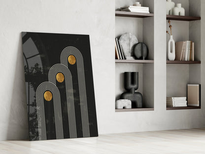 Black glass art print with white line arch patterns and gold circles, leaning against white wall beside wooden shelving unit.