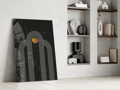 Black glass wall art with white concentric arches and gold circle leaning against white wall beside wooden shelving unit.