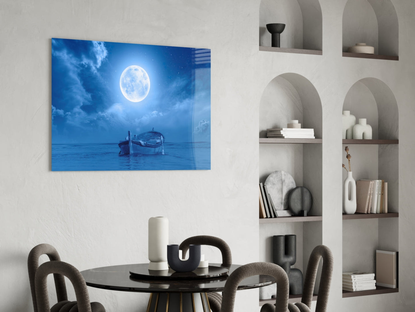 Glass wall art depicting a moonlit ocean scene with a wooden boat, displayed above a round black dining table in a modern interior.