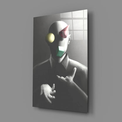 Angled view of glass wall art showing monochromatic sculptural figure with colored geometric shapes on face against gridded window background.