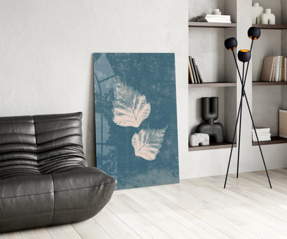 Teal glass wall art with white leaf prints displayed in modern living room next to black leather chair and floor lamp.