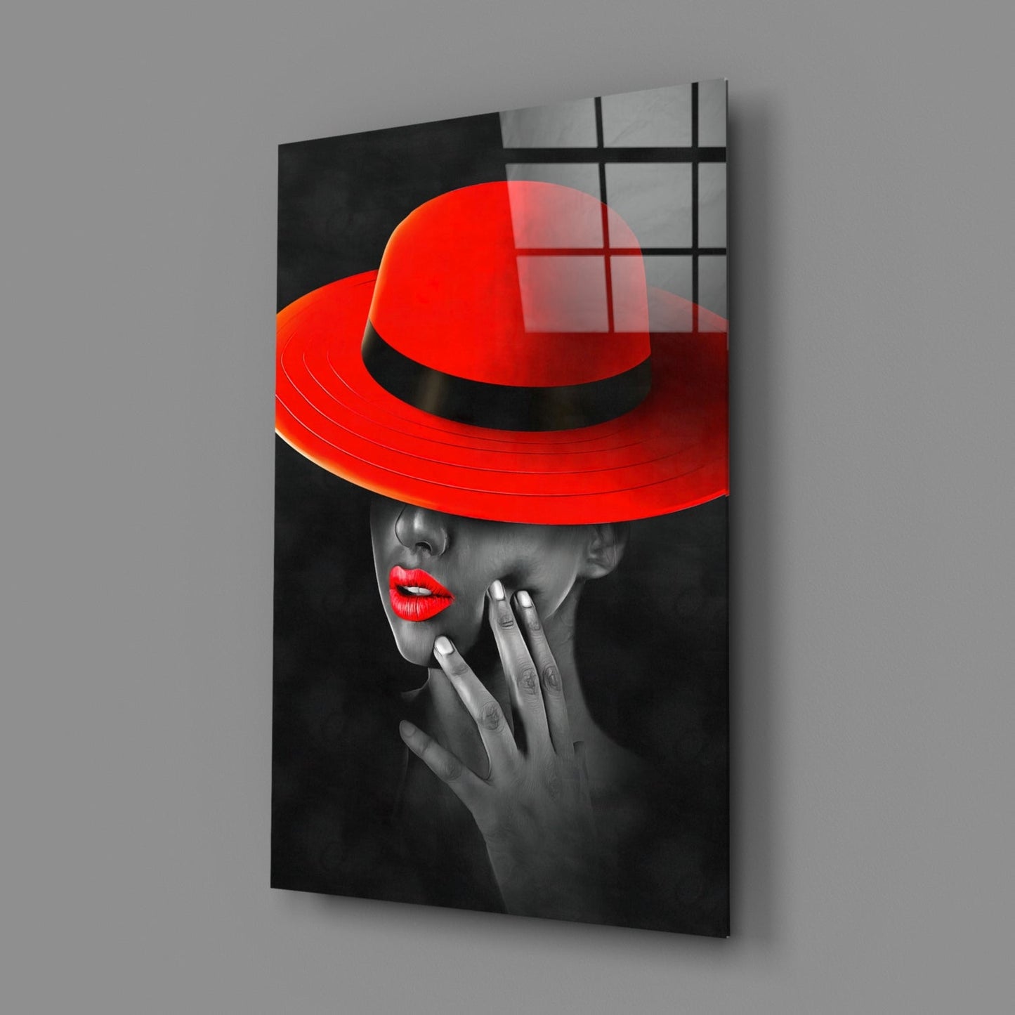 Angled perspective of glass wall art showing woman in red hat and red lips against black and white background, mounted on gray wall.