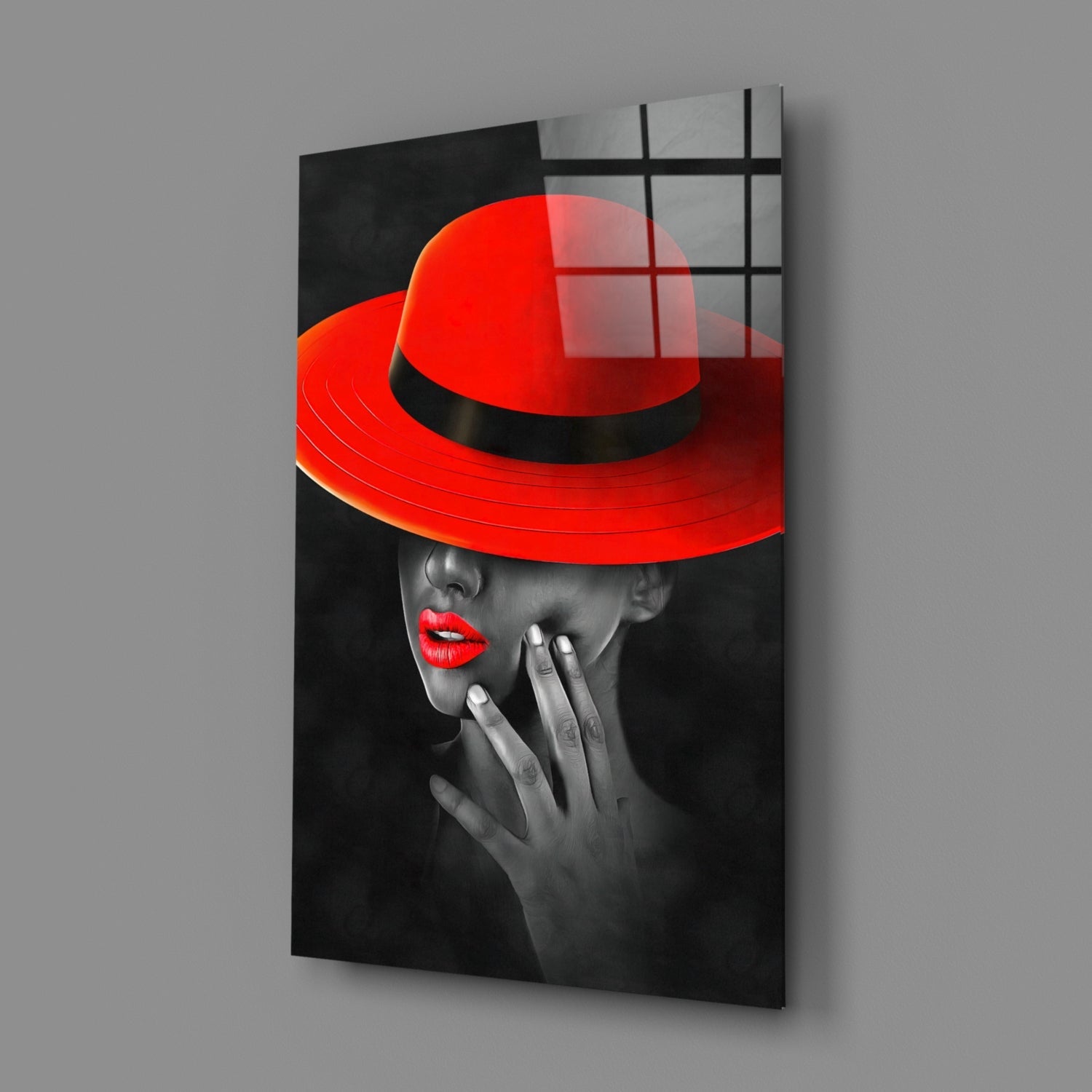Angled perspective of glass wall art showing woman in red hat and red lips against black and white background, mounted on gray wall.