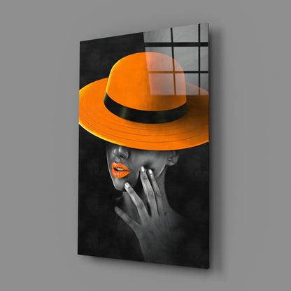 Orange Hat Glass Wall Art displayed at an angle showing a woman in grayscale wearing a vibrant orange wide-brimmed hat and lipstick.