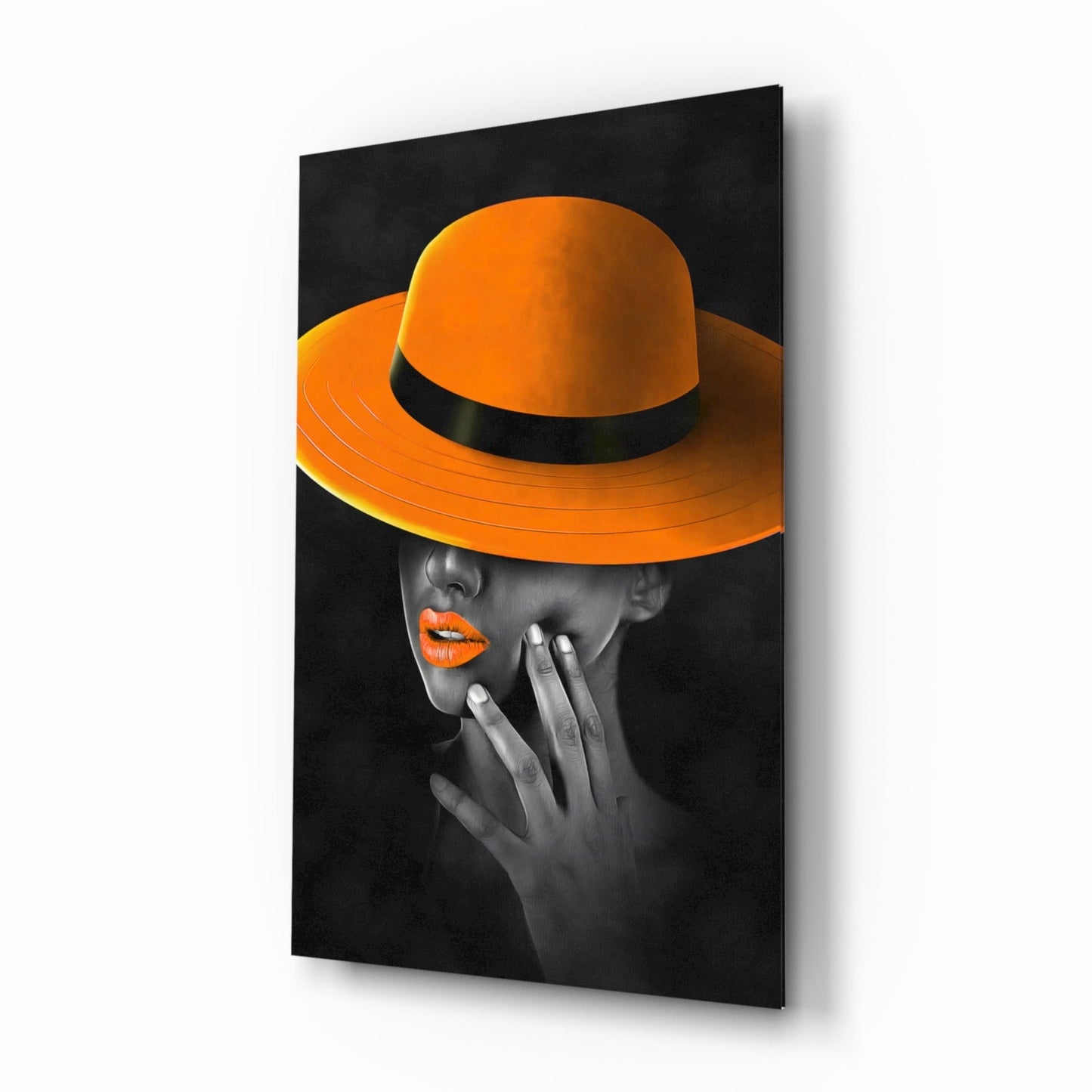 Angled view of glass wall art showing woman in grayscale wearing bright orange wide-brimmed hat with black band and orange lipstick against black background.