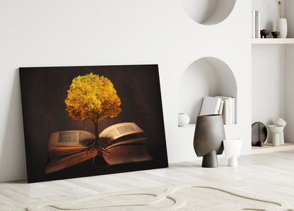 Black glass wall art featuring golden glowing tree growing from open book pages, leaning against white wall beside decorative objects.