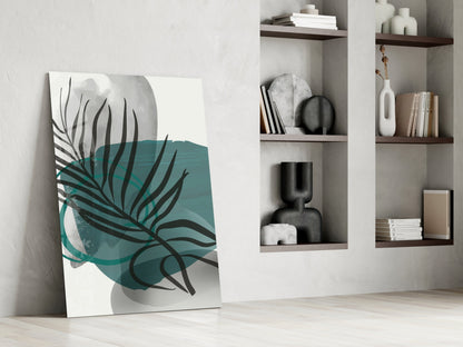 Modern glass wall art with teal and black botanical leaf design leaning against white wall beside shelving.