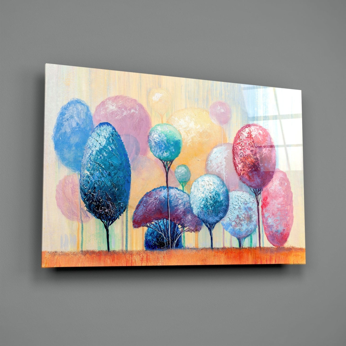 Colorful abstract glass wall art depicting stylized trees with textured oval canopies in blue, pink, yellow, and red against a cream background with orange ground, displayed at an angle on gray wall.