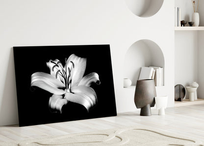 Black and white lily glass wall art leaning against white wall in minimalist interior setting with decorative objects.