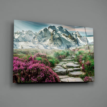 Glass wall art mounted on gray wall showing snow-capped mountains with pink flowering plants lining a stone pathway in foreground.