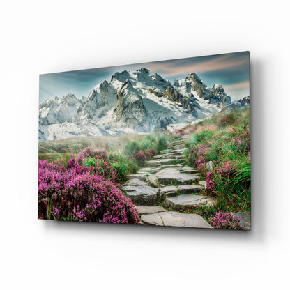 Glass wall art displaying a stone pathway lined with magenta flowers leading toward snow-capped mountains under blue sky.