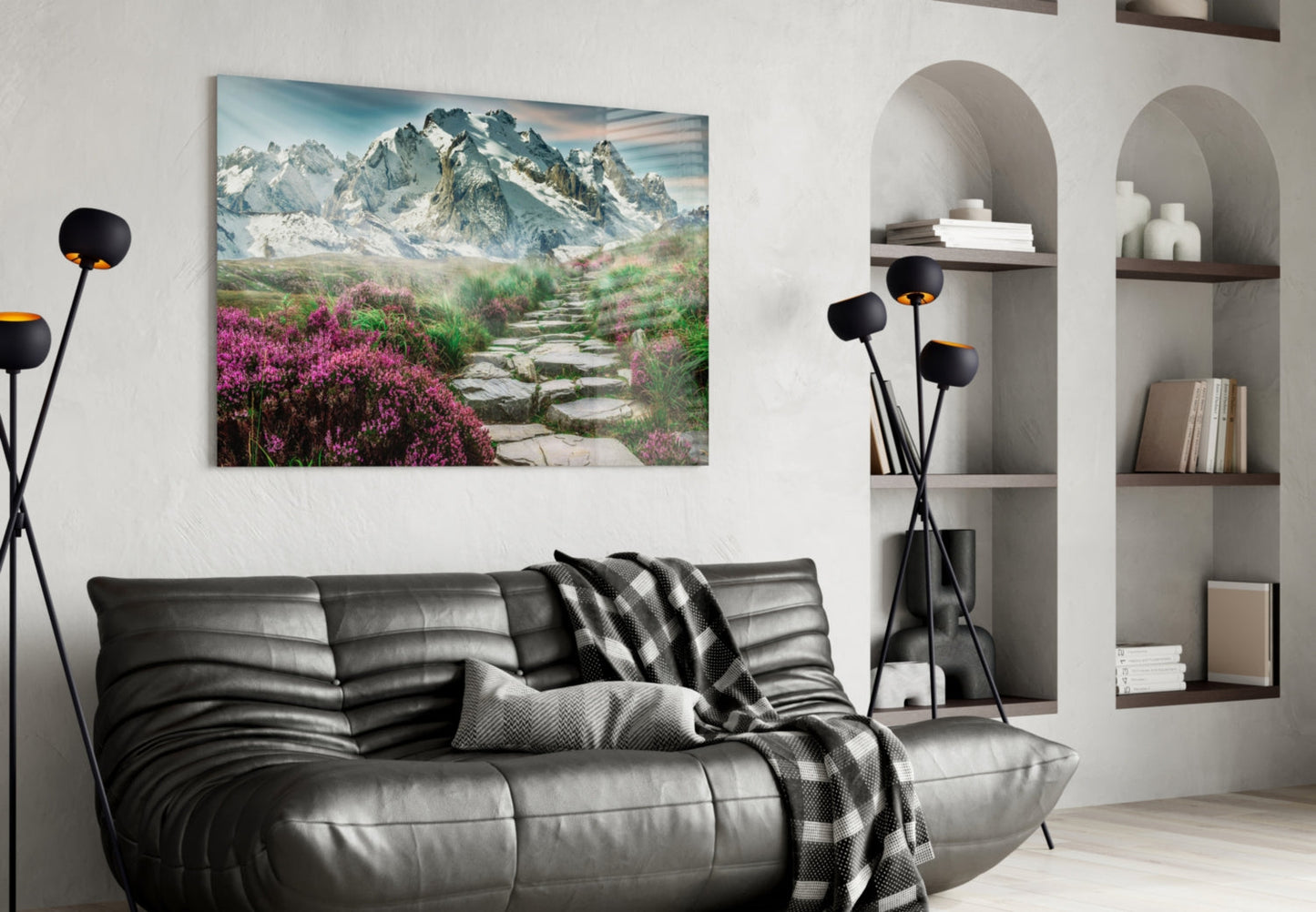 Glass wall art depicting snow-capped mountains and pink flowering meadow with stone pathway, displayed above black leather sofa in modern living room.