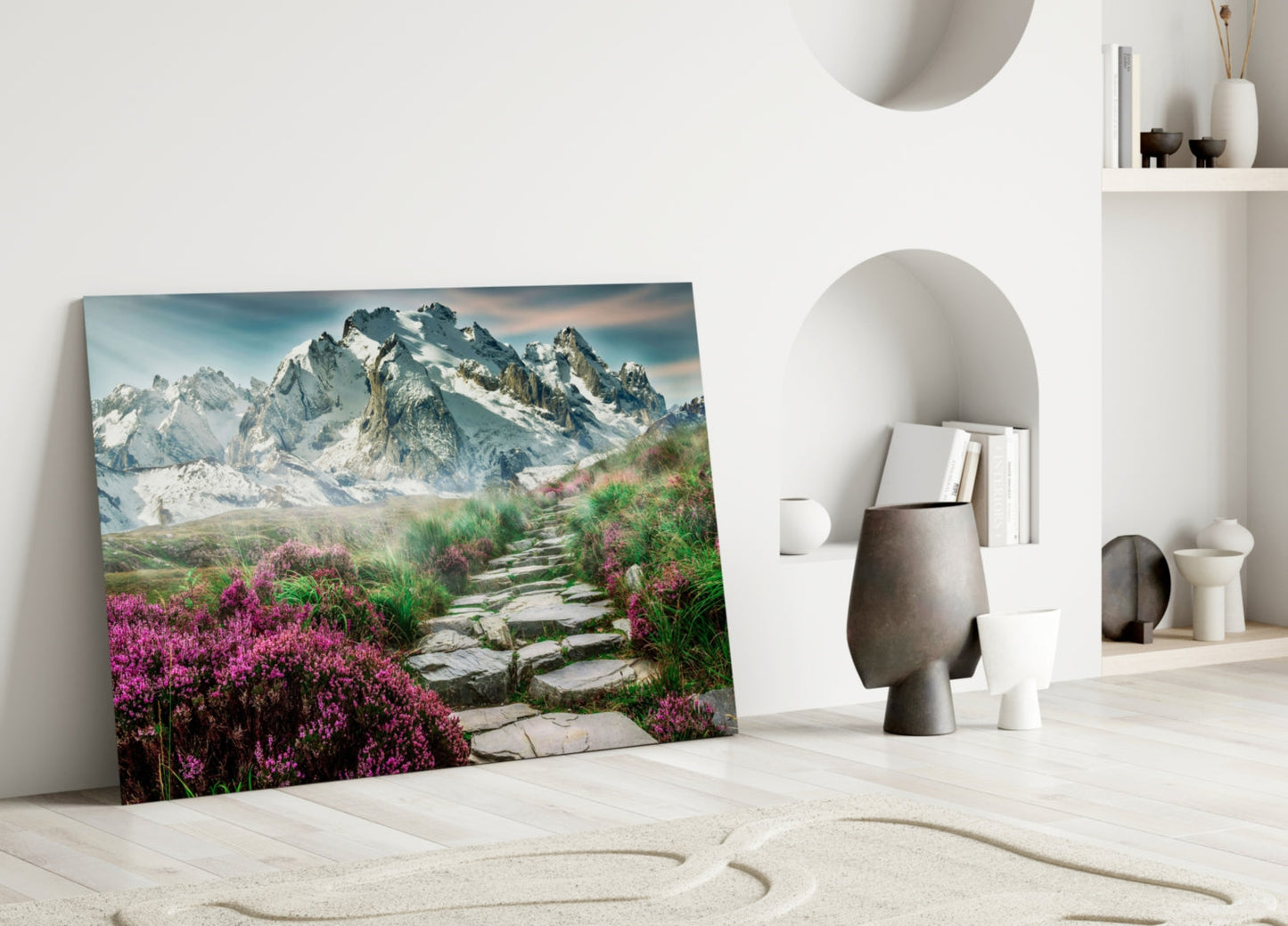 Glass wall art leaning against white wall displays snow-capped mountains with purple wildflowers and stone pathway in foreground.