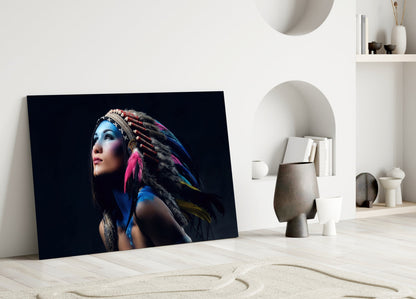 Glass wall art panel leaning against white wall displays woman with blue face paint and colorful feathered headdress against black background.
