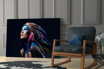 Glass wall art displaying a woman with blue face paint and colorful feathered headdress against black background, displayed in modern living room setting.