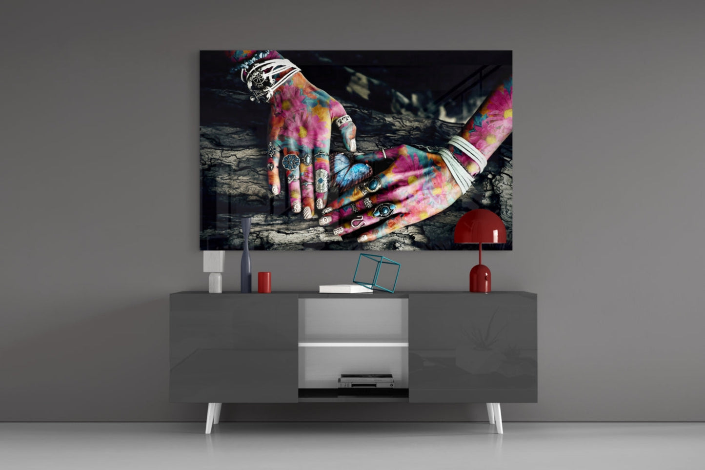 Colorful tattooed hands displayed on glass art mounted above gray media console with red lamp and decorative accessories.