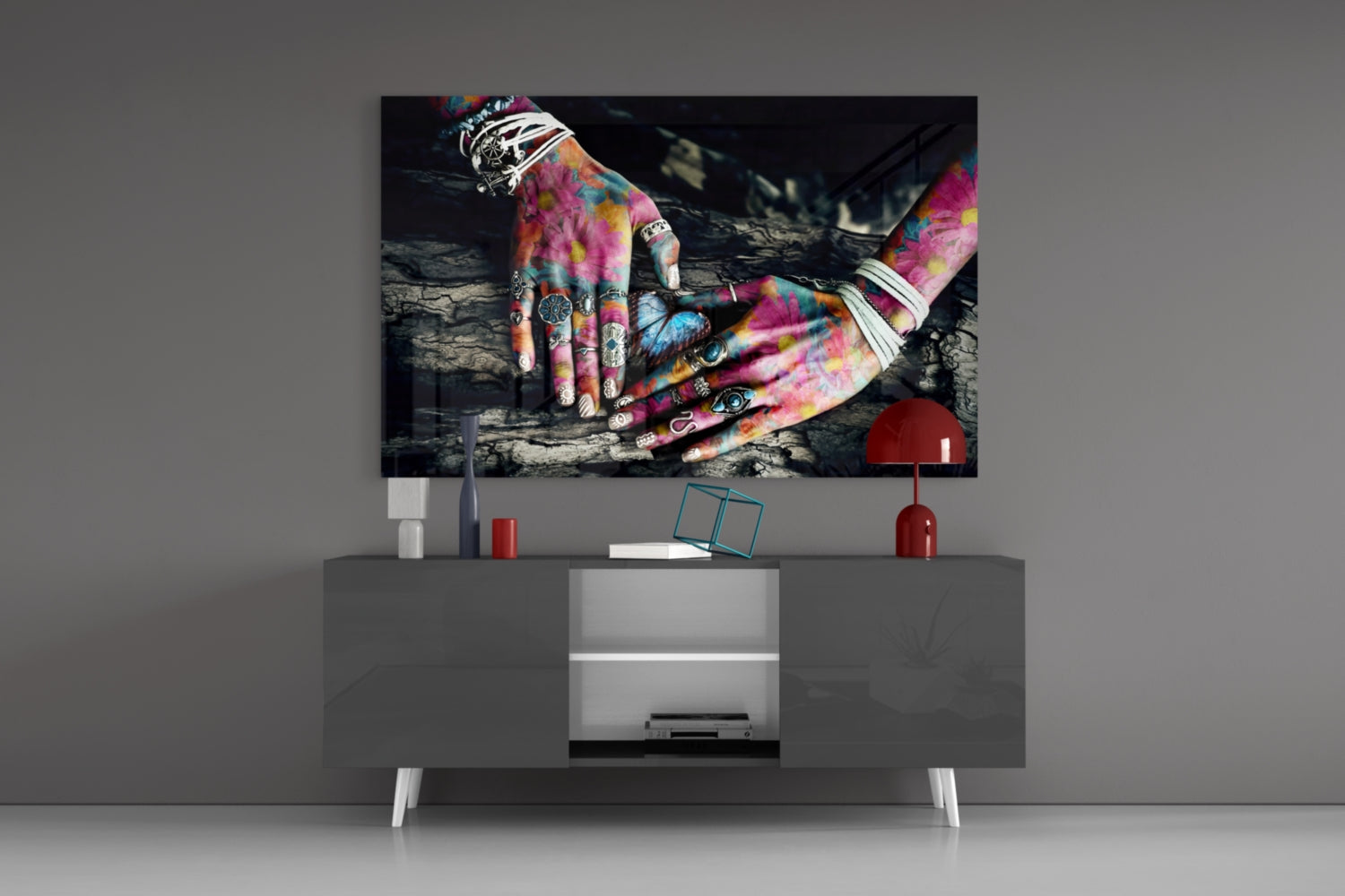 Colorful tattooed hands displayed on glass art mounted above gray media console with red lamp and decorative accessories.