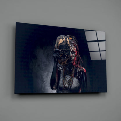 Glass wall art mounted on gray wall showing masked figure in ornate gold and black mask with red fabric and beaded necklaces against dark background.