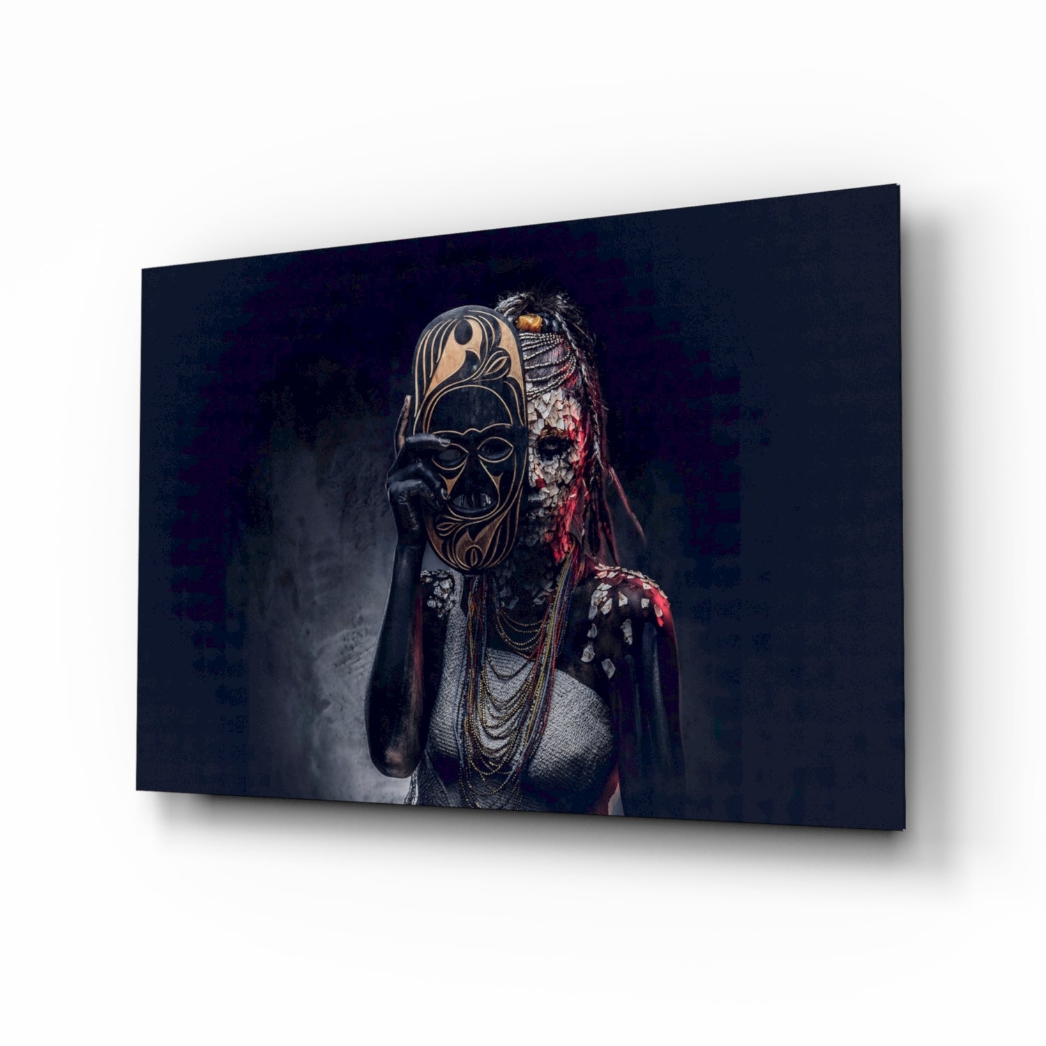 Angled perspective of glass wall art displaying figure in ornate tribal mask and beaded necklaces against dark background.