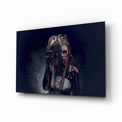 Angled perspective of glass wall art displaying figure in ornate tribal mask and beaded necklaces against dark background.